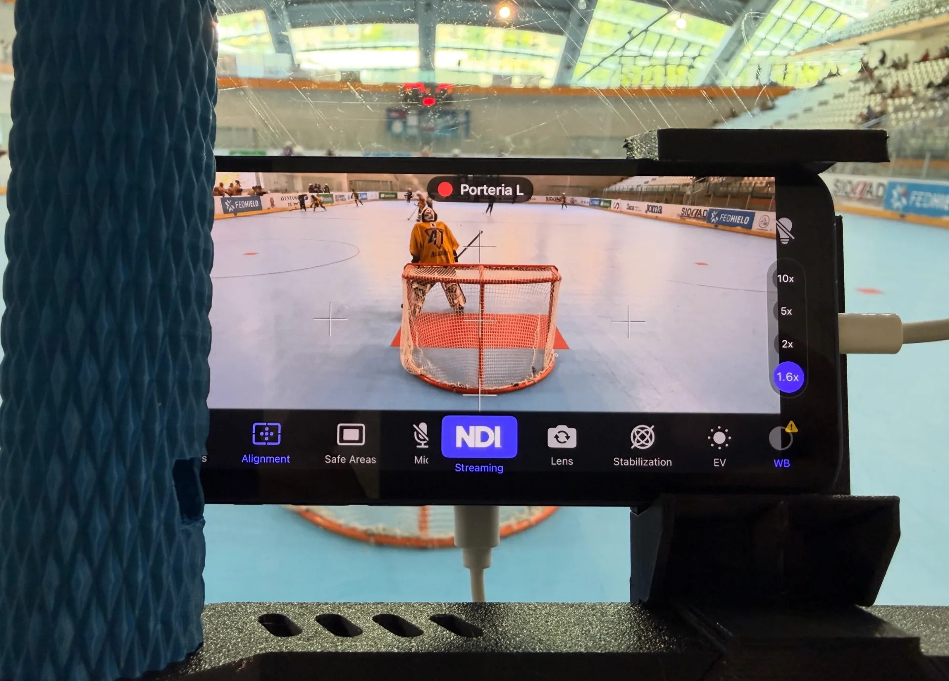 Cámara iPhone con app NDI enfocando la portería desde detrás del cristal durante una retransmisión de hockey línea en pabellón